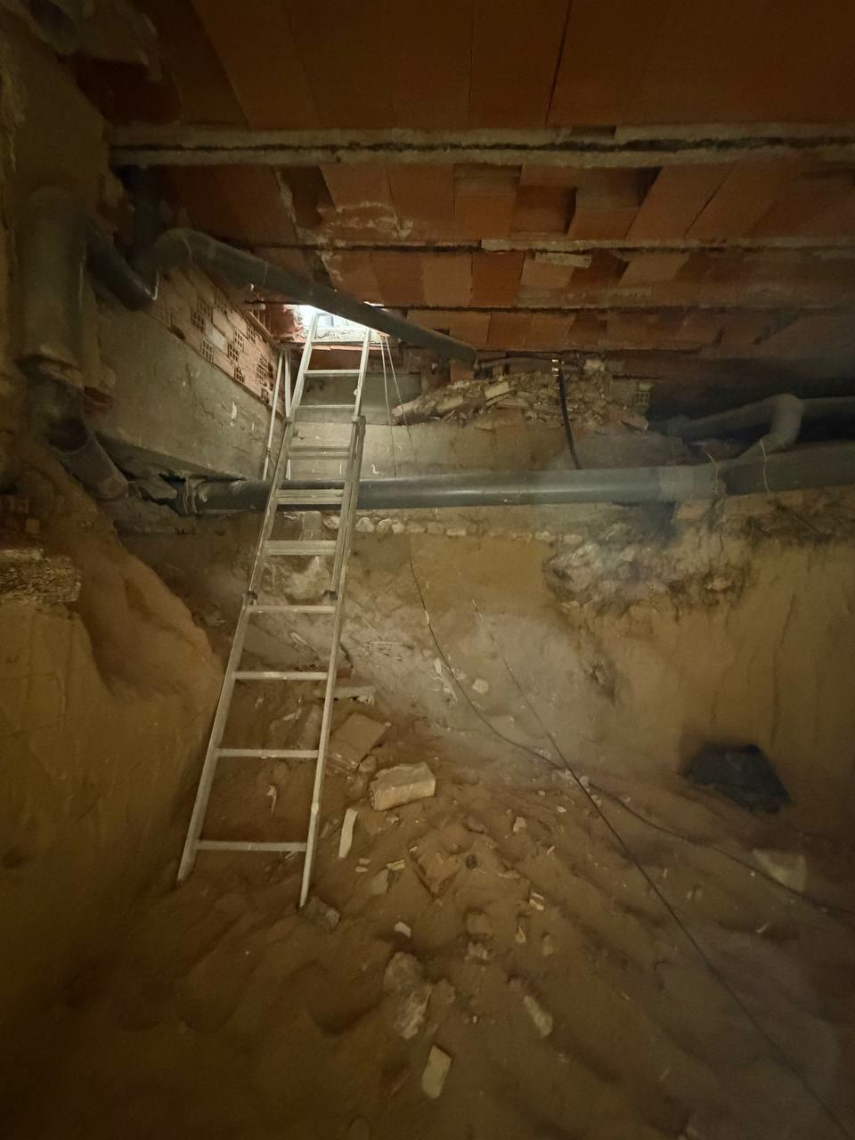 Excavación bajo la vivienda con una escalera de mano apoyada, tuberías bajo el techo y escombros sobre la tierra